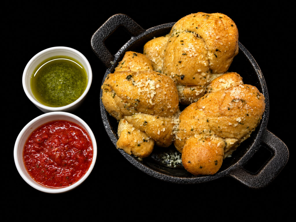 Buttery garlic knots displayed in a cast iron dish with dipping cups filled with pesto and marinara