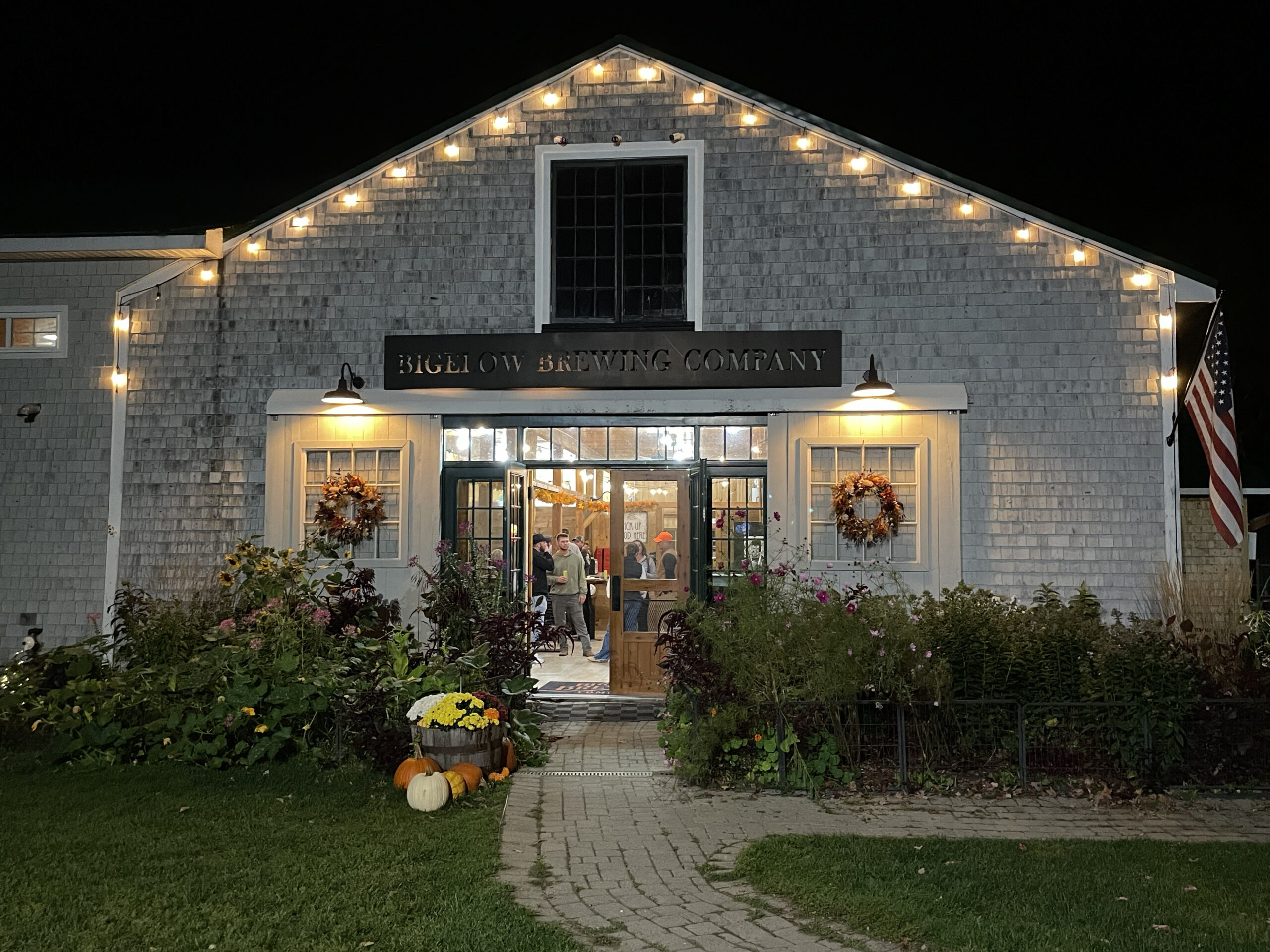 A string of lights line the barns roof on a Saturday evening and people can be seen in the light though the door. Wreaths at the entryway also are shown under light creating a very nice ambiance.