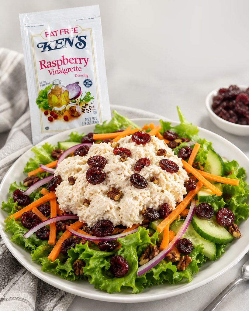 Scoops of chicken salad over mixed greens with cucumber slices, shredded carrots, red onion, dried cranberries, and toasted pecans in a clear takeout container, with a packet of raspberry vinaigrette tucked into the open lid.