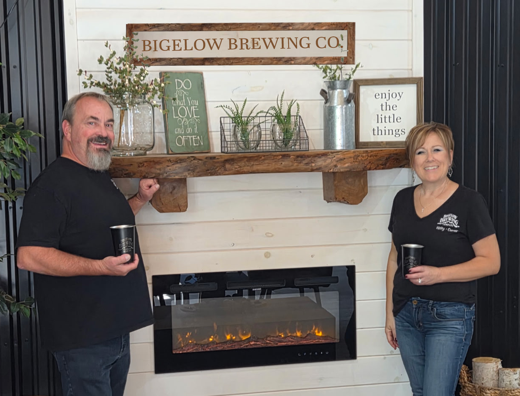 two people stand in front of a cozy fireplace with a mantle holding flowers and signage.