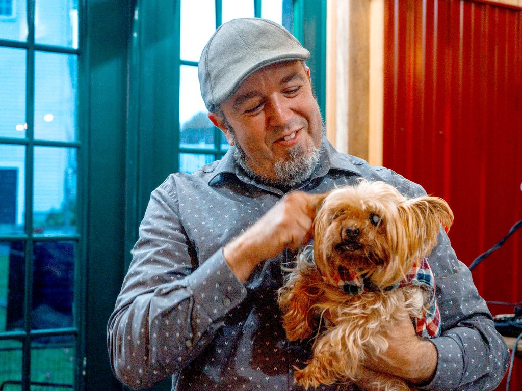 Ty Brown stand inside the green doors with windows at Bigleow Brewion holding a a small dog wearing a sweater.